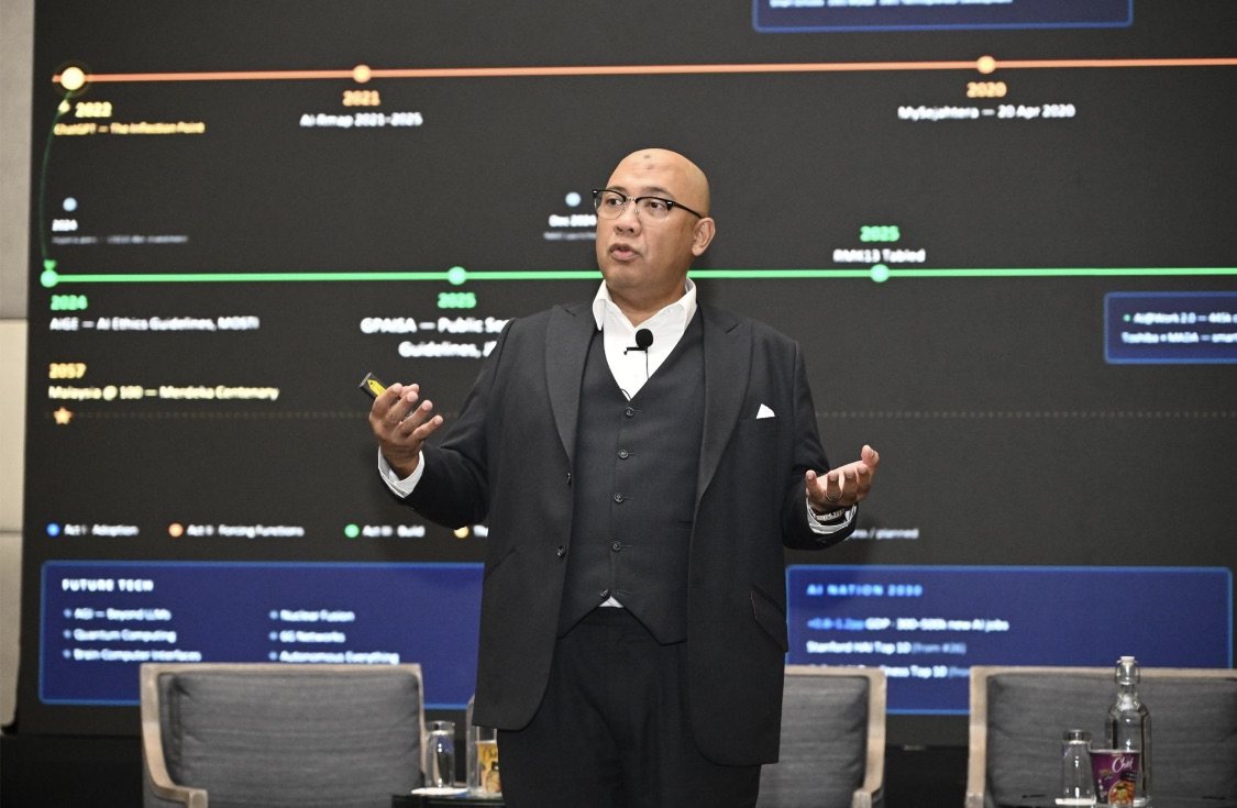 Reza Ali on stage at the FutureCIO Malaysia Conference, mid-sentence with his hands raised, in front of a screen showing a timeline of Malaysia's AI and governance milestones.