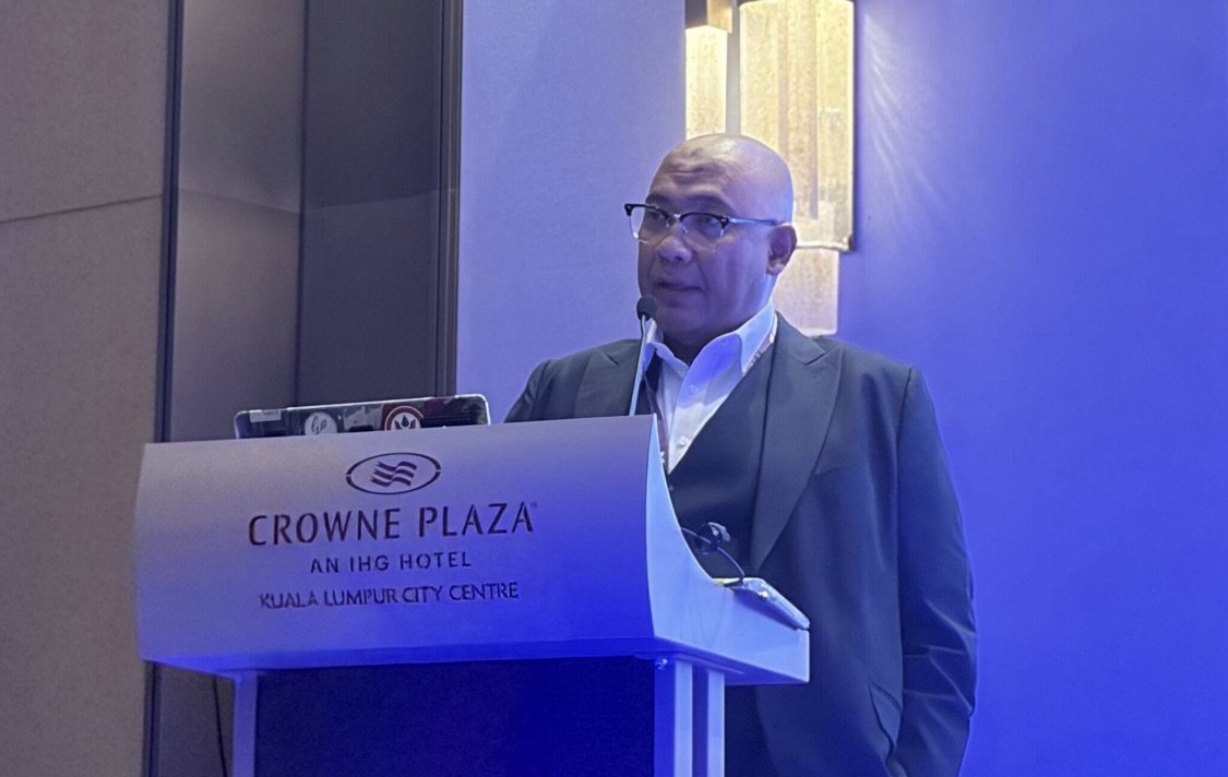 Reza Ali speaking at a Crowne Plaza Kuala Lumpur City Centre lectern at the GovXcellence Summit Malaysia 2026, wearing a dark suit over a white shirt.