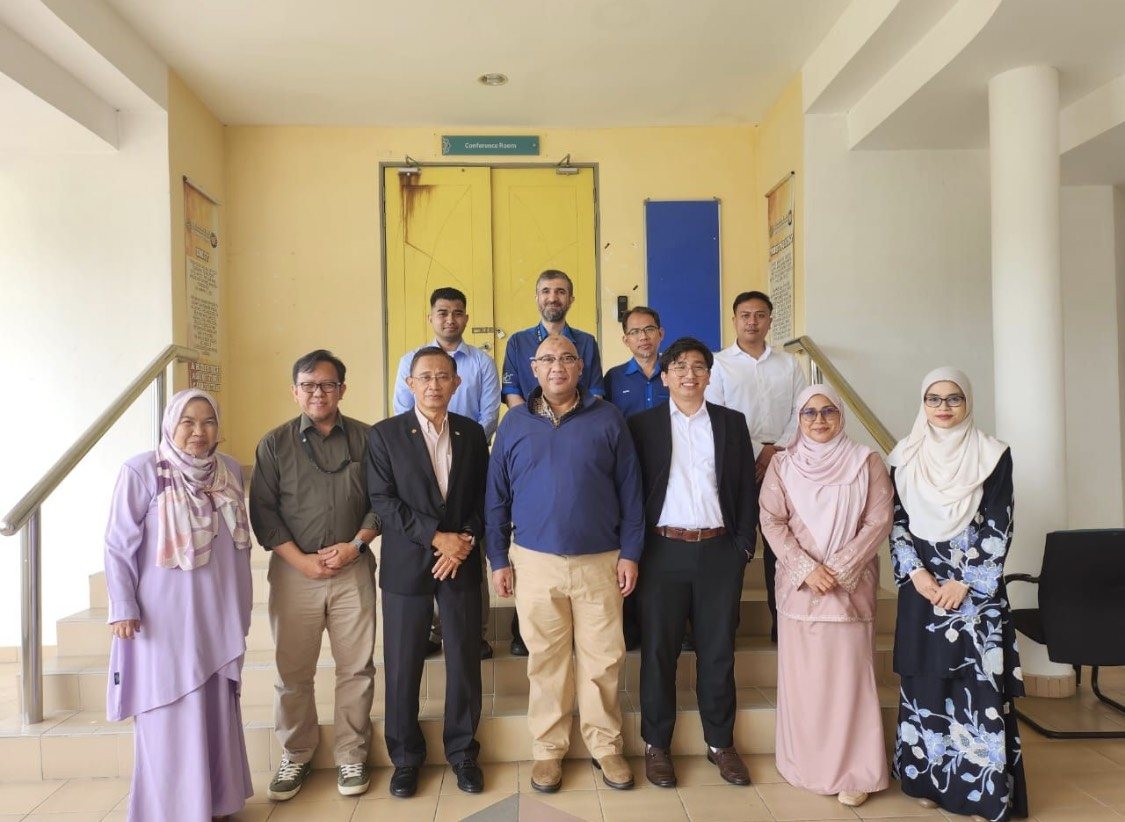 Eleven people standing on the steps of a building at IIUM for a group photograph, in front of yellow doors beneath a Conference Room sign. Reza Ali stands at the centre front in a blue quarter-zip top.