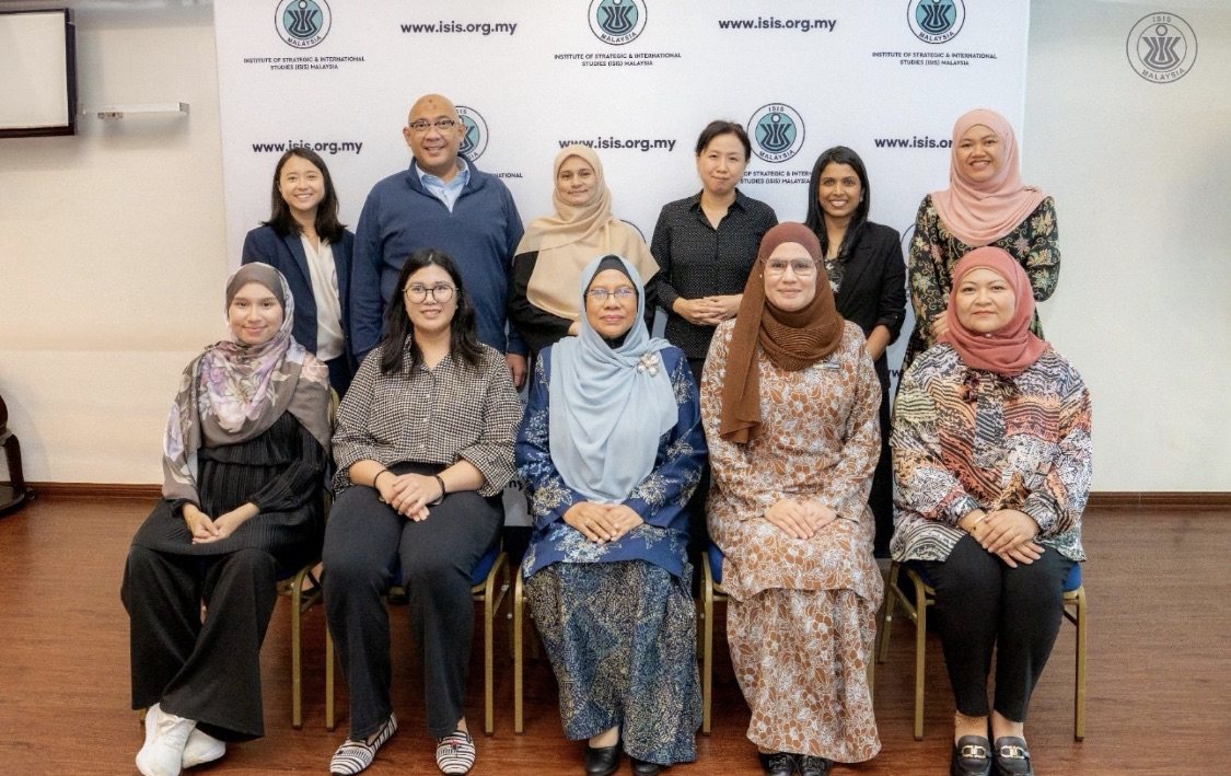 Ten people arranged in two rows in front of an ISIS Malaysia backdrop for a group photograph. Reza Ali stands second from the left in the back row, in a navy quarter-zip top.