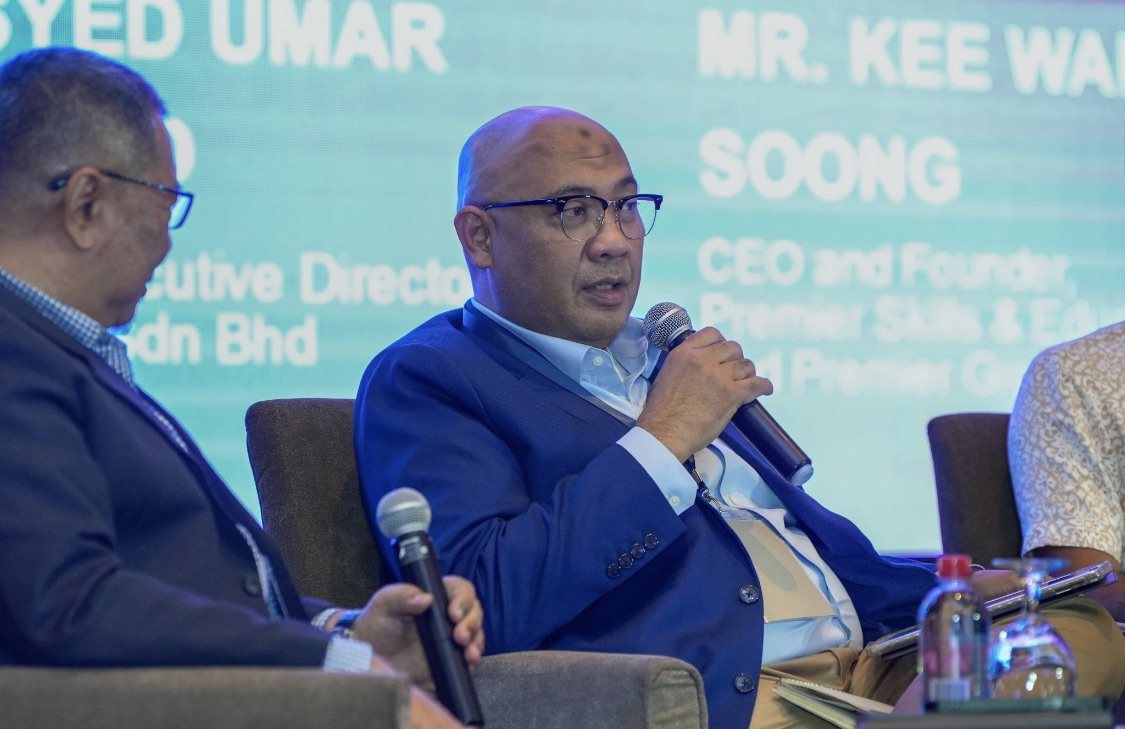 Reza Ali speaking into a handheld microphone during the roundtable, in a blue blazer over a light blue shirt, with fellow panellists seated either side of him.