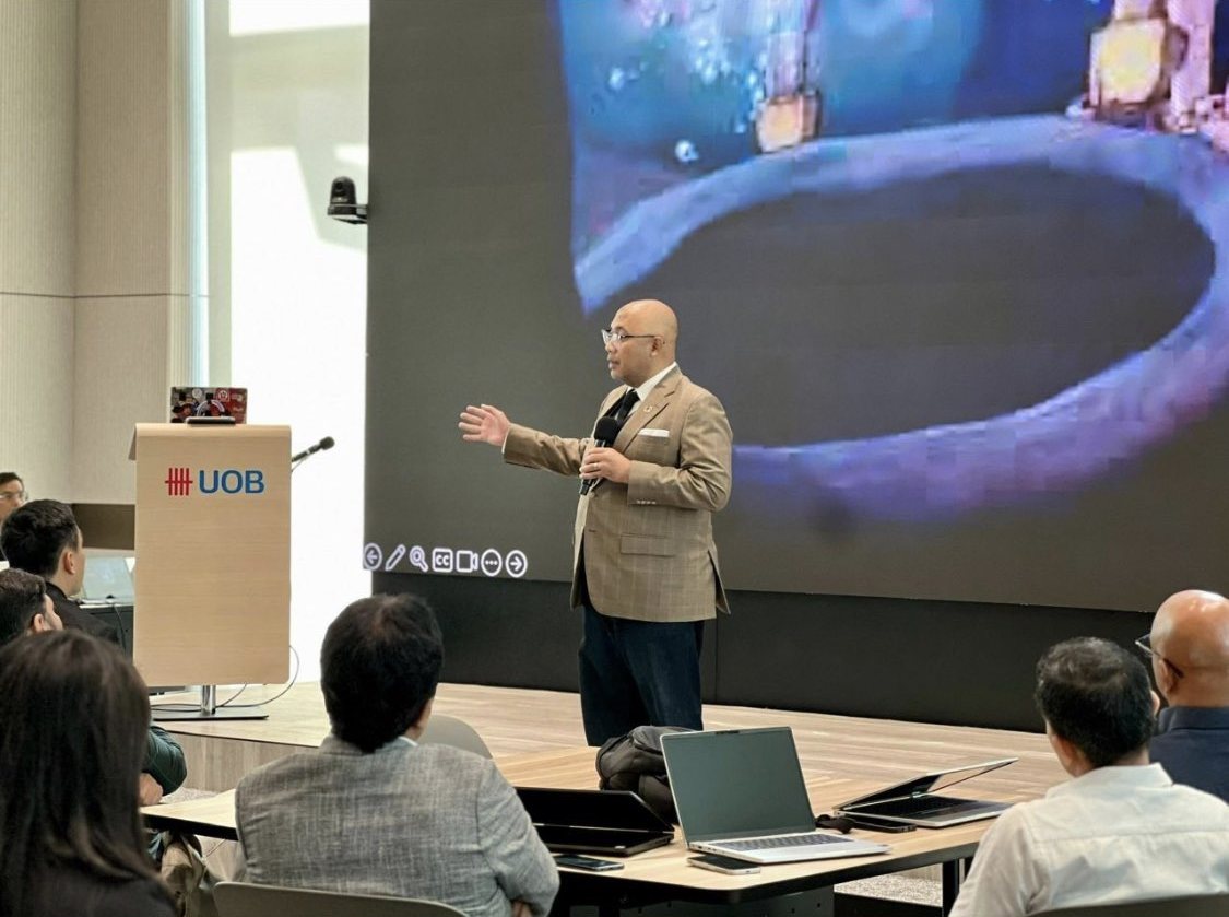 Reza Ali speaking on stage in a tan blazer and dark tie, gesturing with one hand beside a UOB lectern, with an audience of business people seated at tables below him.