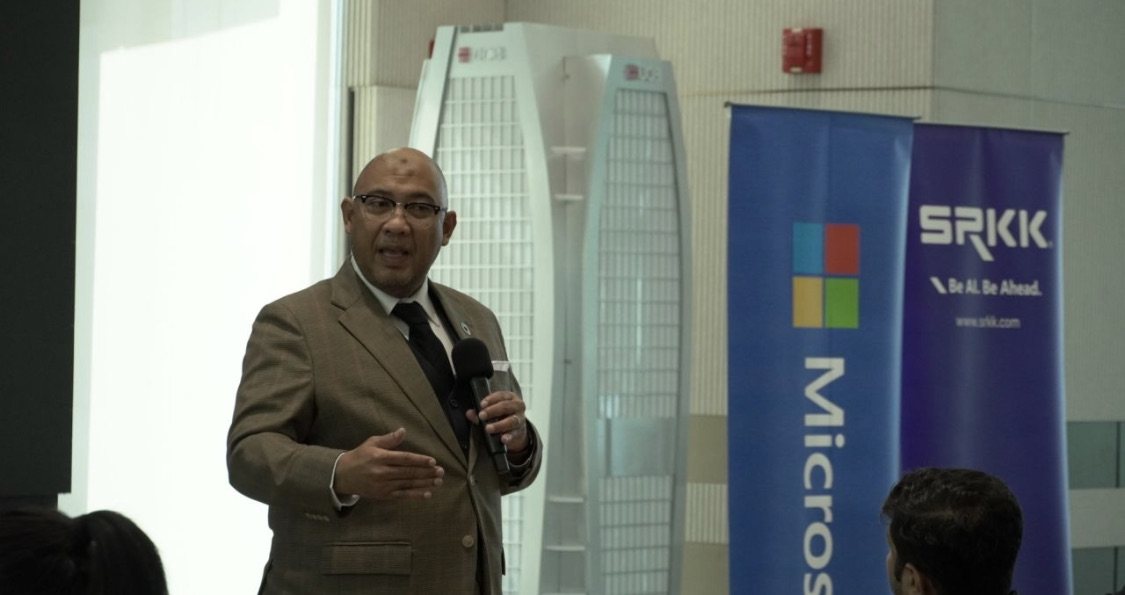 Reza Ali speaking into a microphone in a tan blazer and dark tie, in front of Microsoft and SRKK banners.