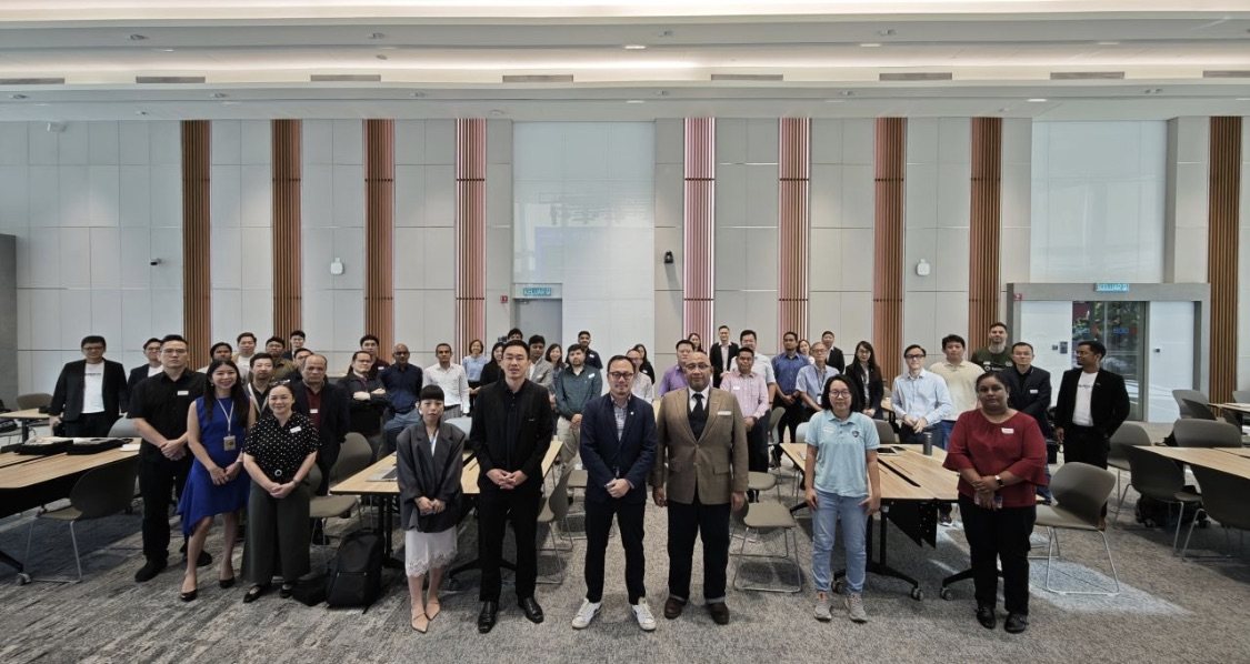 Around thirty participants standing in rows for a group photograph in a conference hall, among tables and chairs, in front of wood-panelled walls.