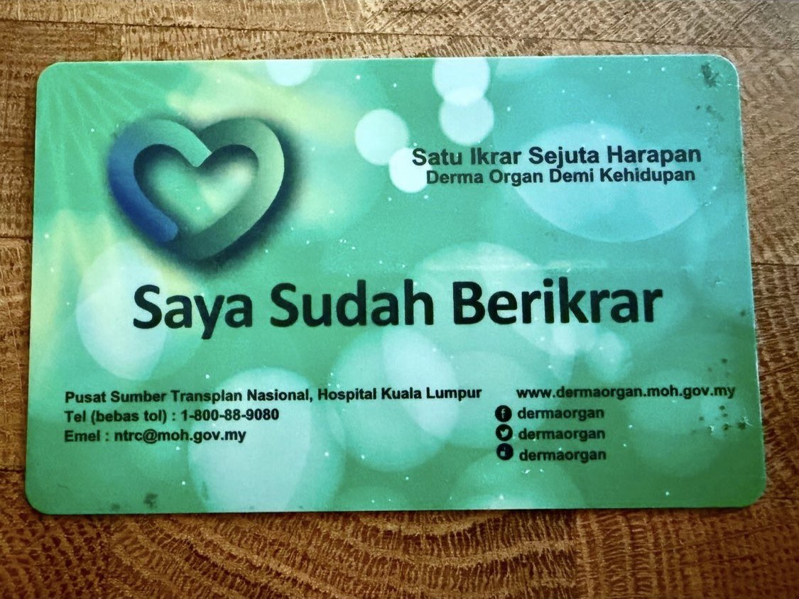 A Malaysian organ donor pledge card lying on a wooden surface. The green card carries a blue heart logo and the words 'Saya Sudah Berikrar' above the National Transplant Resource Centre's telephone and email details.